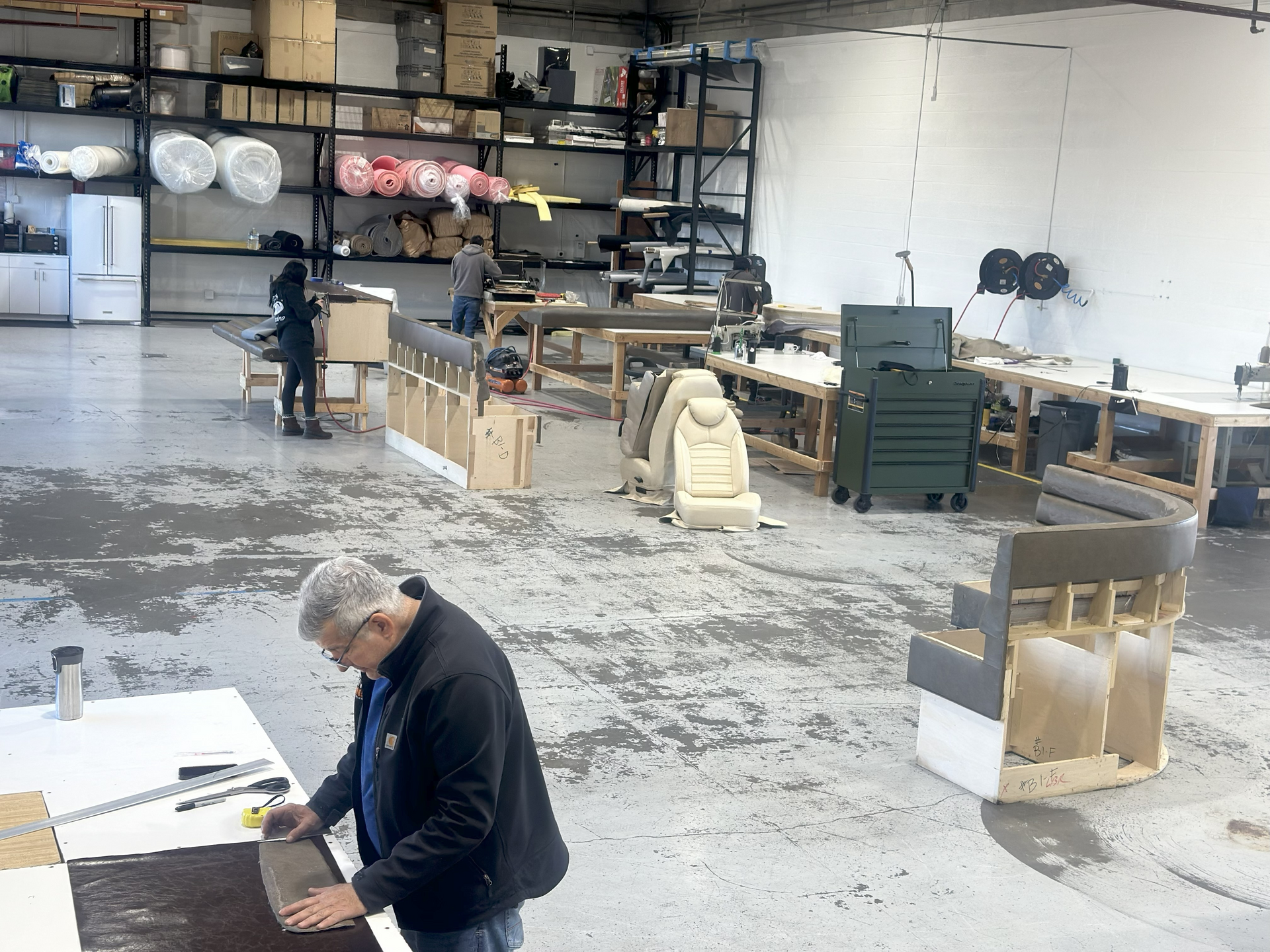 People working in a workshop; cutting, assembling materials. Interior shot, tools, equipment, and materials visible.