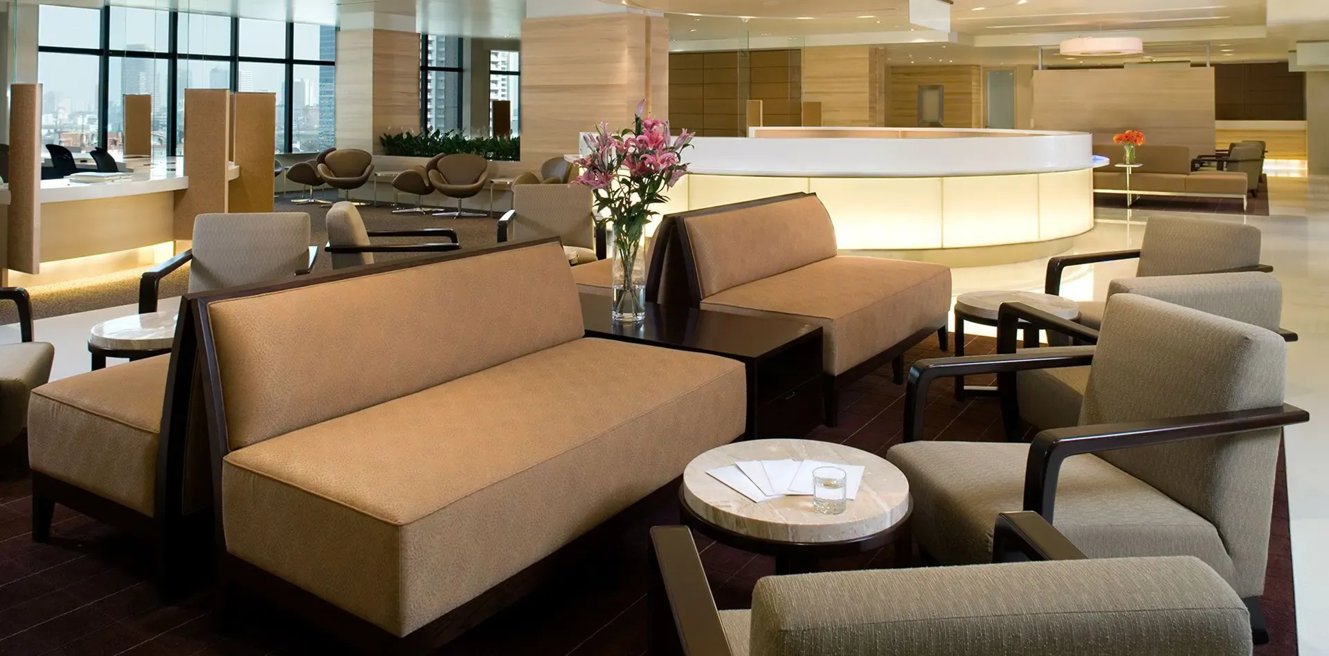 Lobby with beige sofas, chairs, small tables, and a reception desk. A vase of flowers sits on a table.
