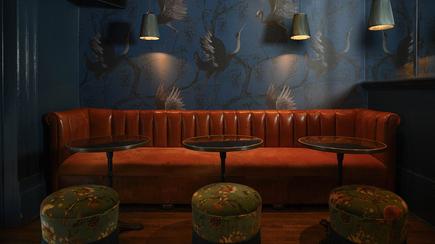 Lounge with orange leather couch, small black tables, green patterned ottomans, and blue floral wallpaper.