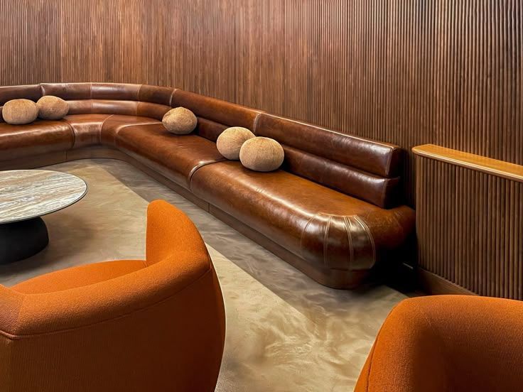 Brown leather sectional sofa with orange chairs in a wood-paneled room; modern interior.