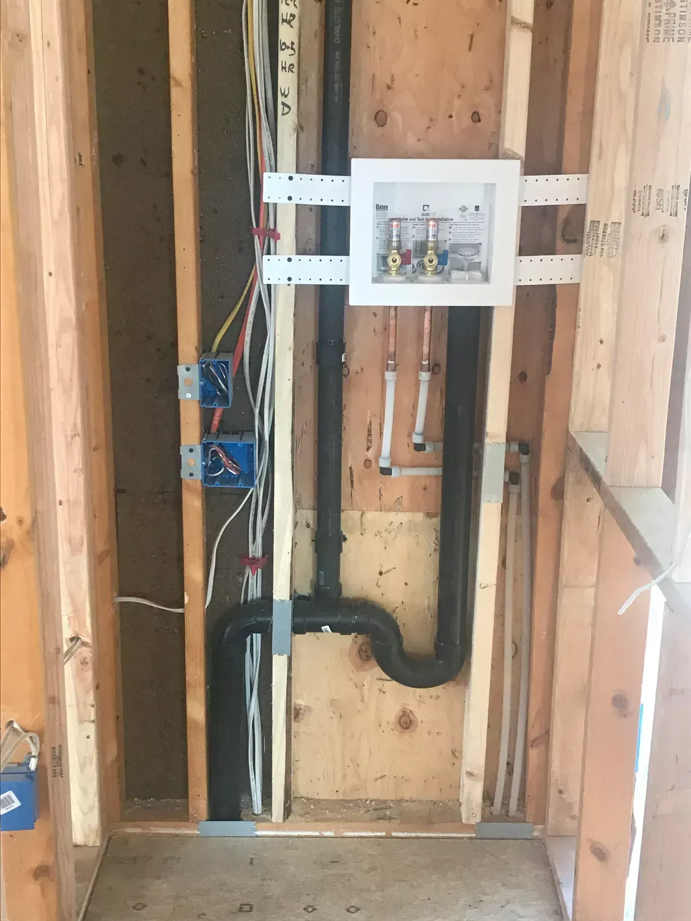 Framed wall with plumbing and electrical fixtures. Black drain pipes, white water box, blue electrical boxes.