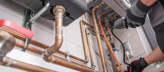 A plumber works on copper pipes connected to a heating system. The setting is indoors, with white tile.