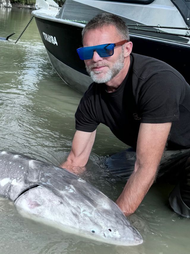 Man kneeling beside a large fish in shallow water, with a boat in the background.