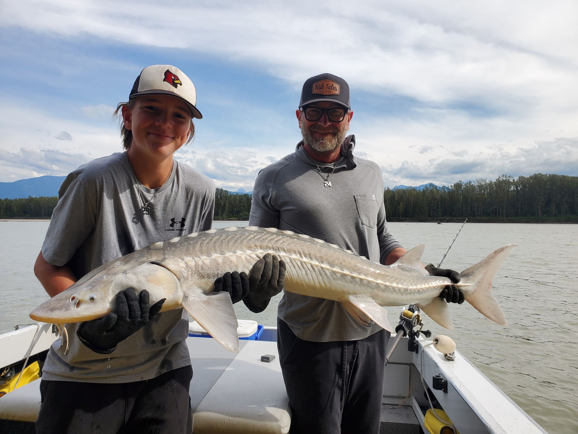 Two anglers on a boat hold a large silver fish on a lake with mountains in the background.