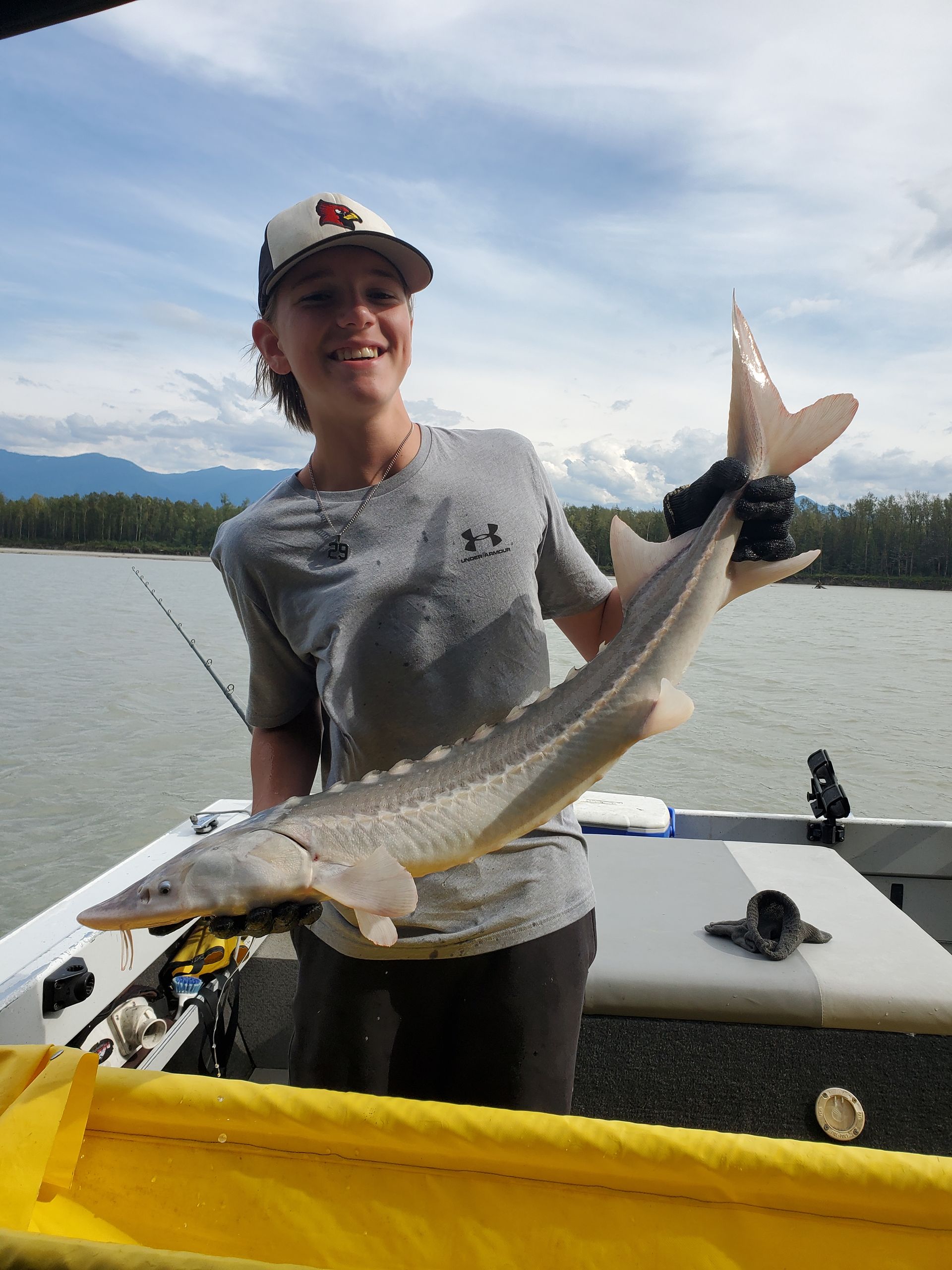 Person smiling and holding a large fish on a boat by a calm lake, with mountains in the background
