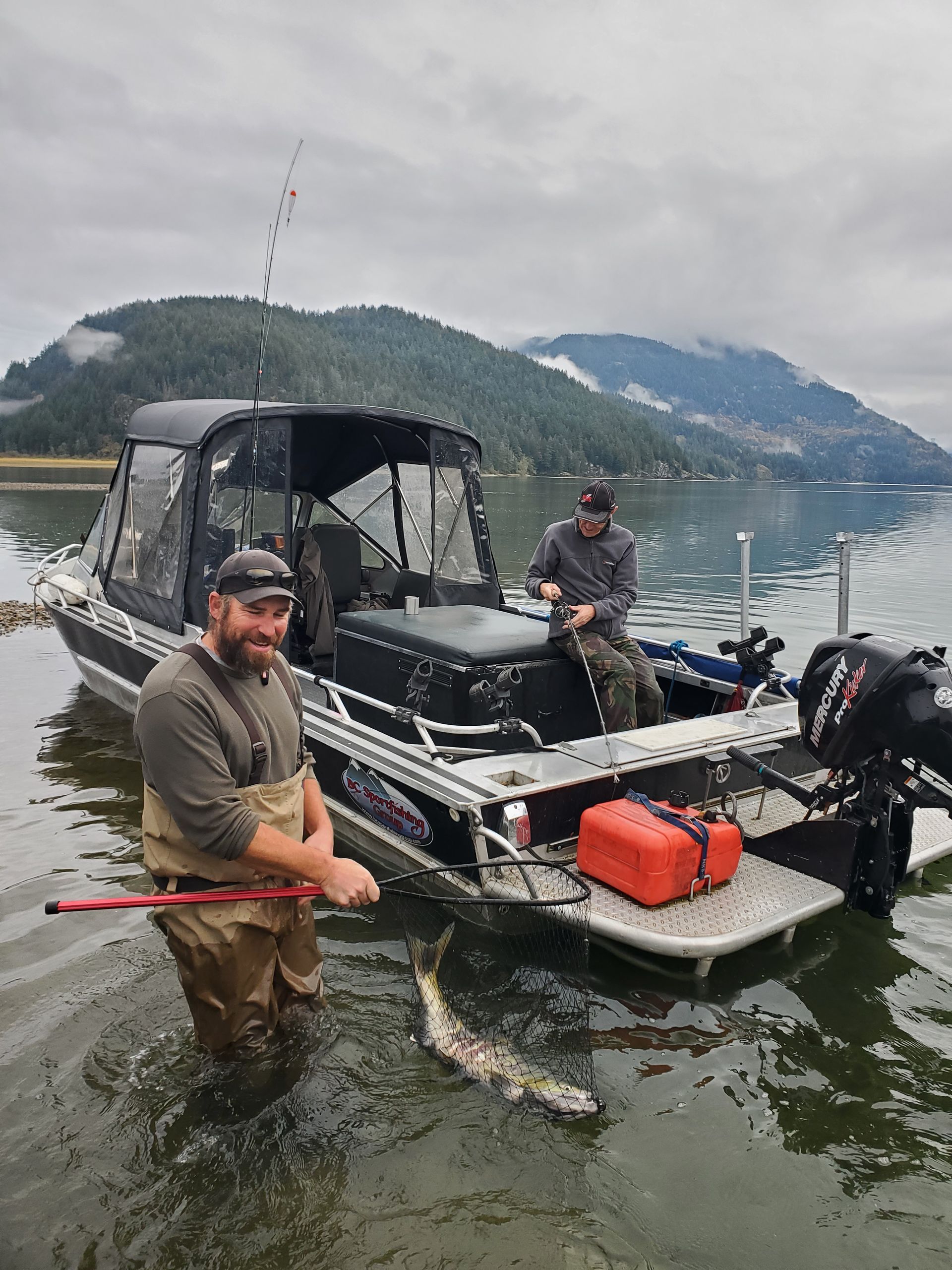 Two men on a small fishing boat by a mountainous lakeshore, one holding a large fish in the water.