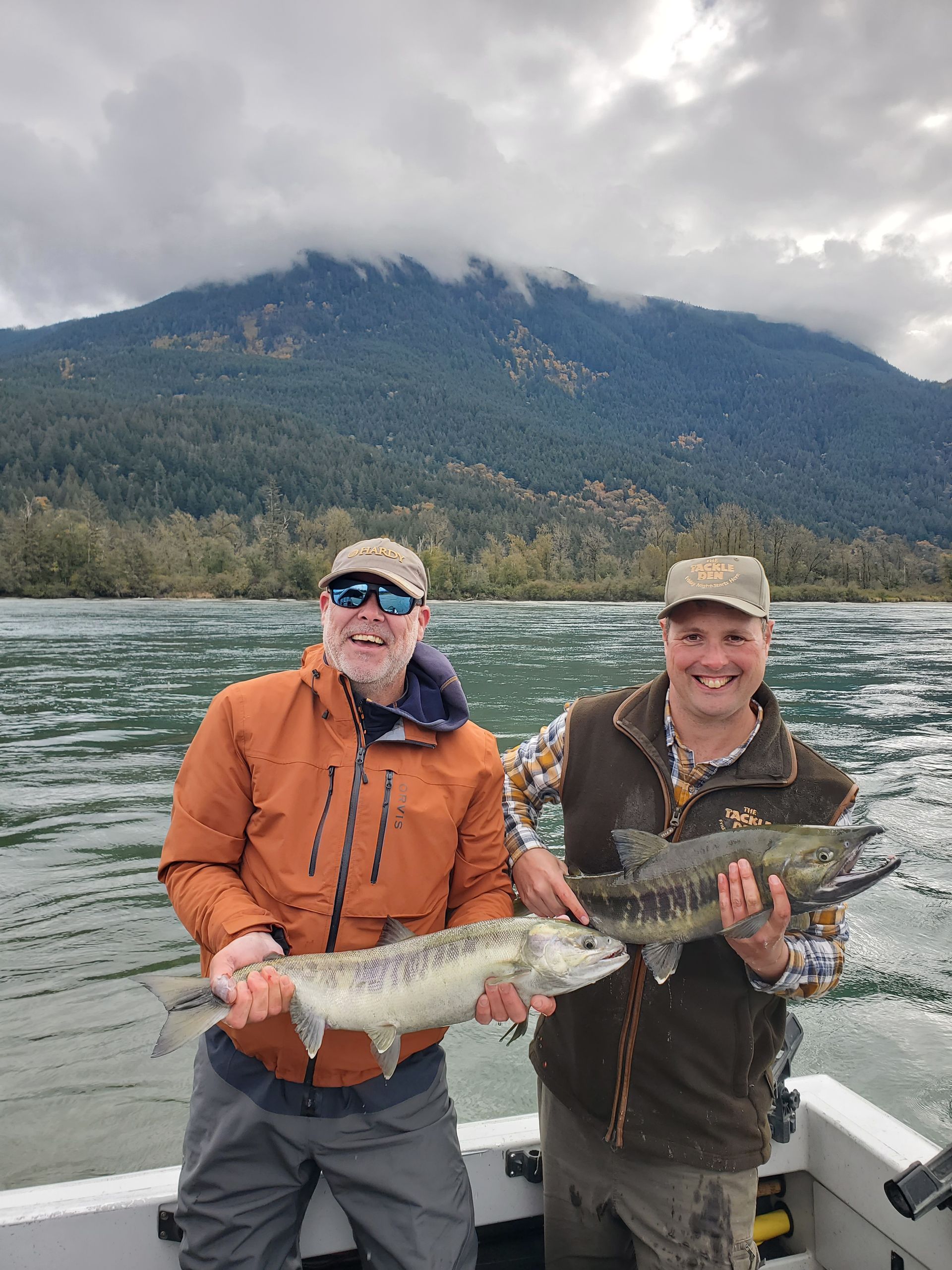 Two anglers on a boat hold large fish on a lake with forested mountains in the background.