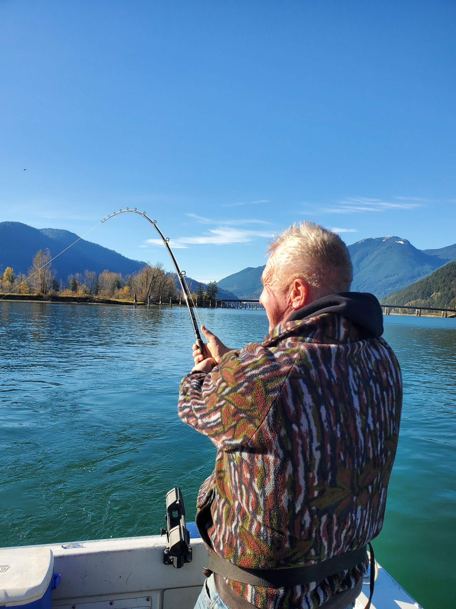 Person fishing from a boat on a lake with mountains and blue sky in the background