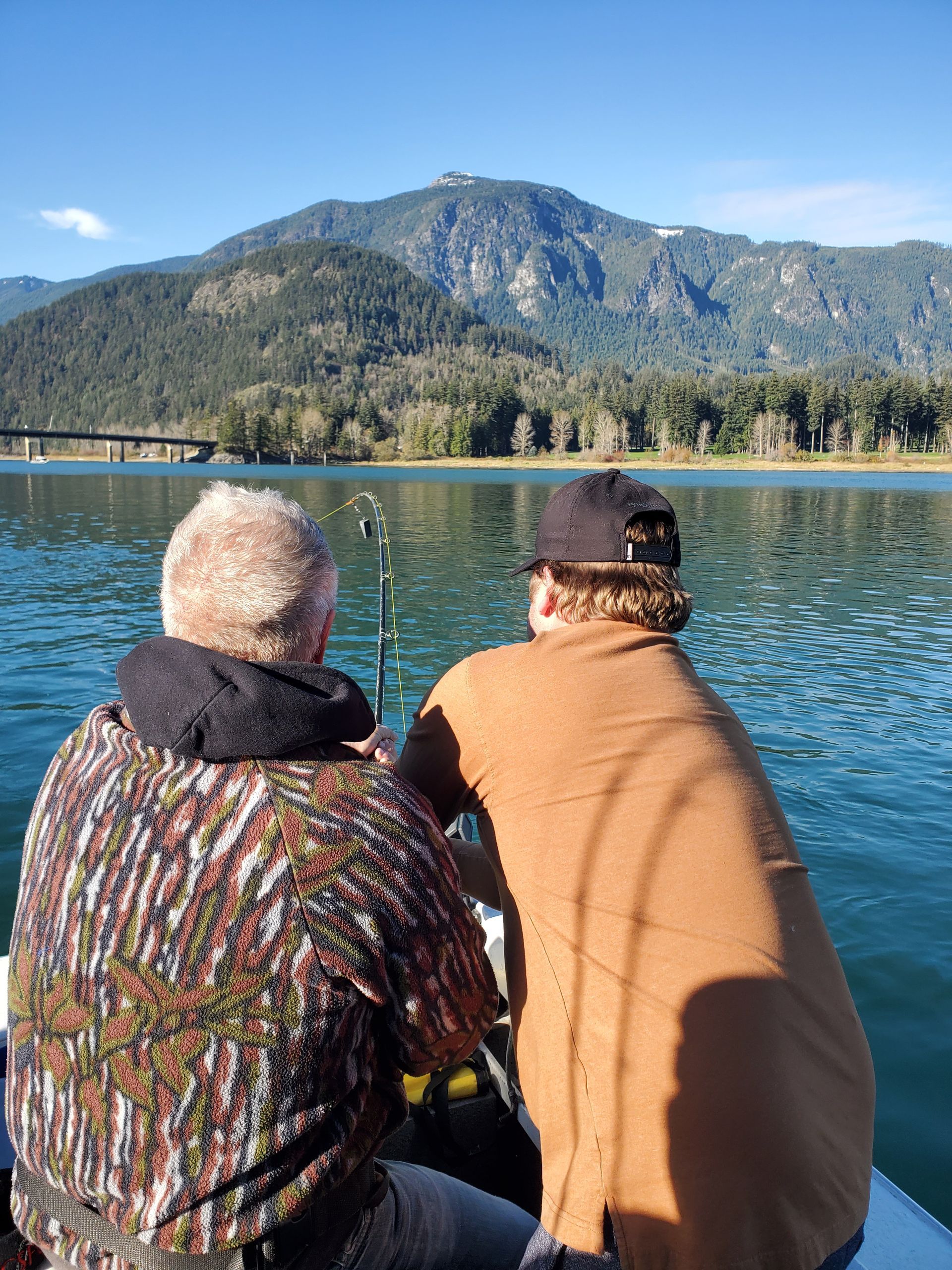 Two people sit by a blue lake facing forested mountains under a clear sky.