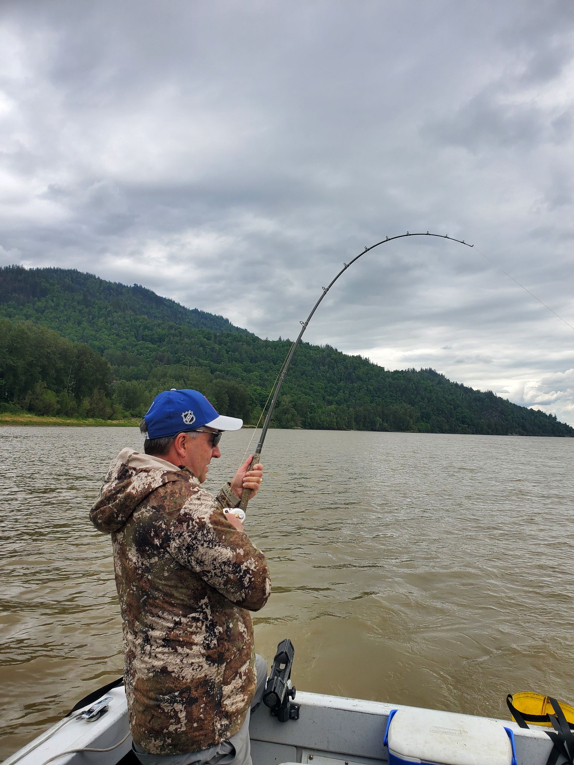 Angler in a blue cap reels in a fish from a boat on a lake with forested hills under cloudy skies