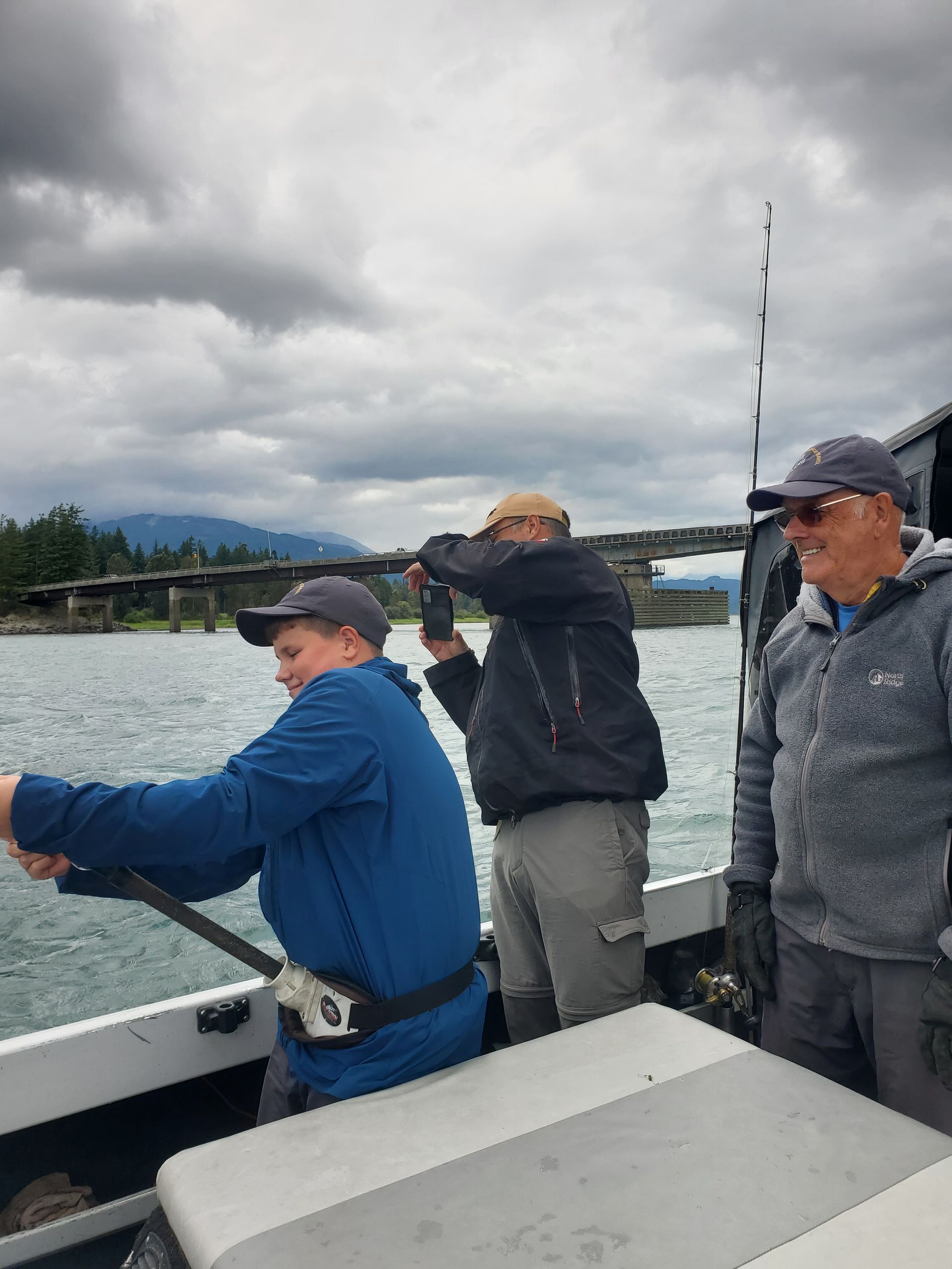 Three people on a boat fishing on a cloudy lake, one casting a line.