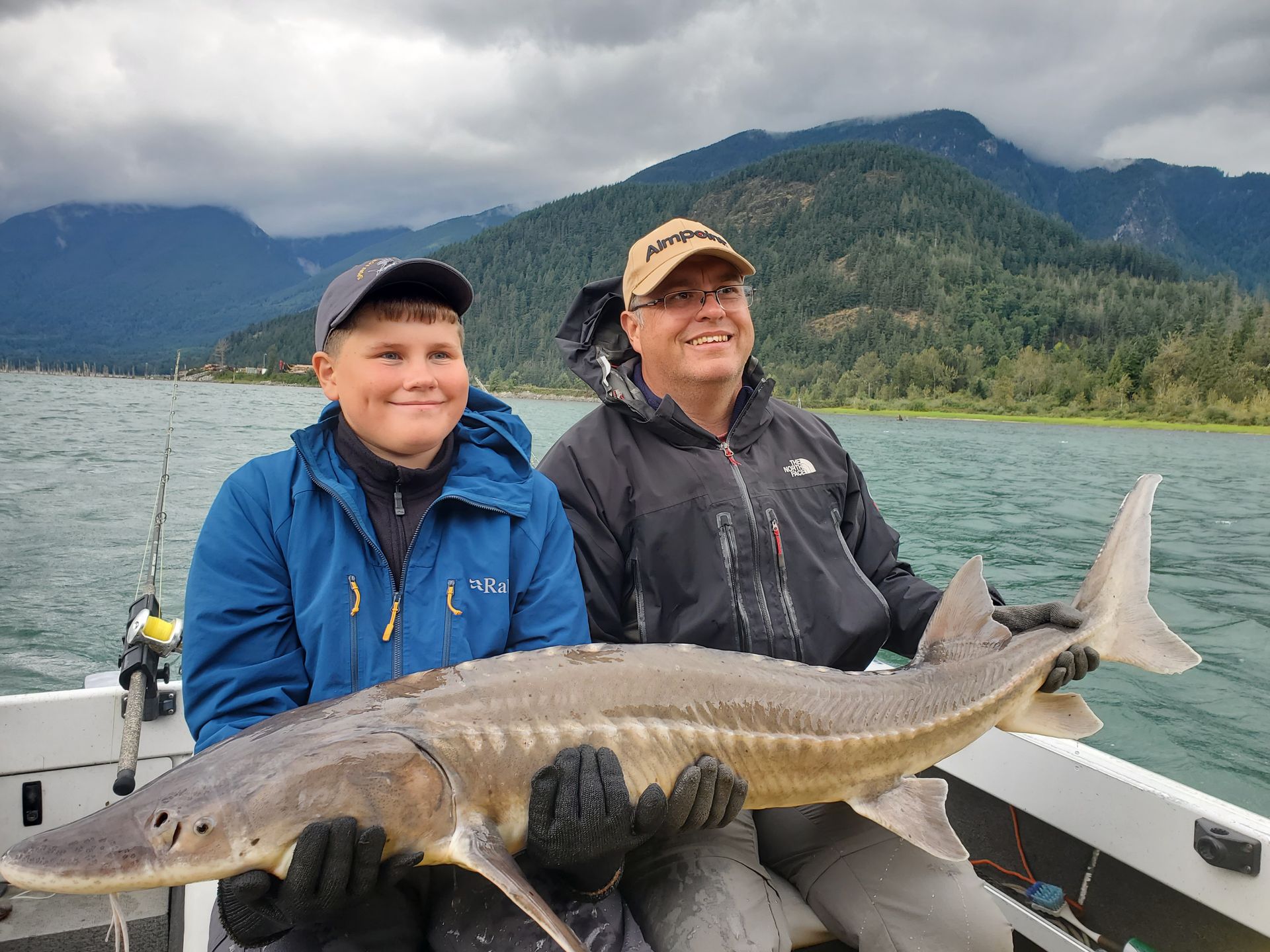 Two people on a boat hold a large fish with mountains and water in the background.