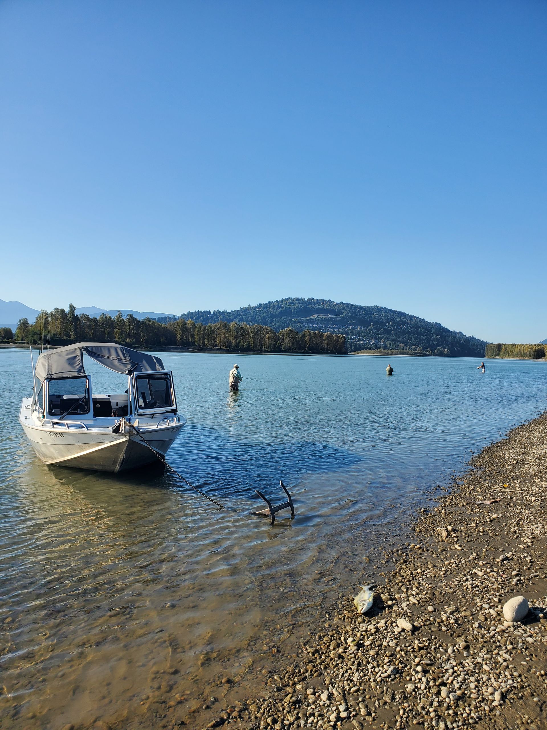 Small boat near a rocky lakeshore with swimmers and a forested hill under a clear blue sky