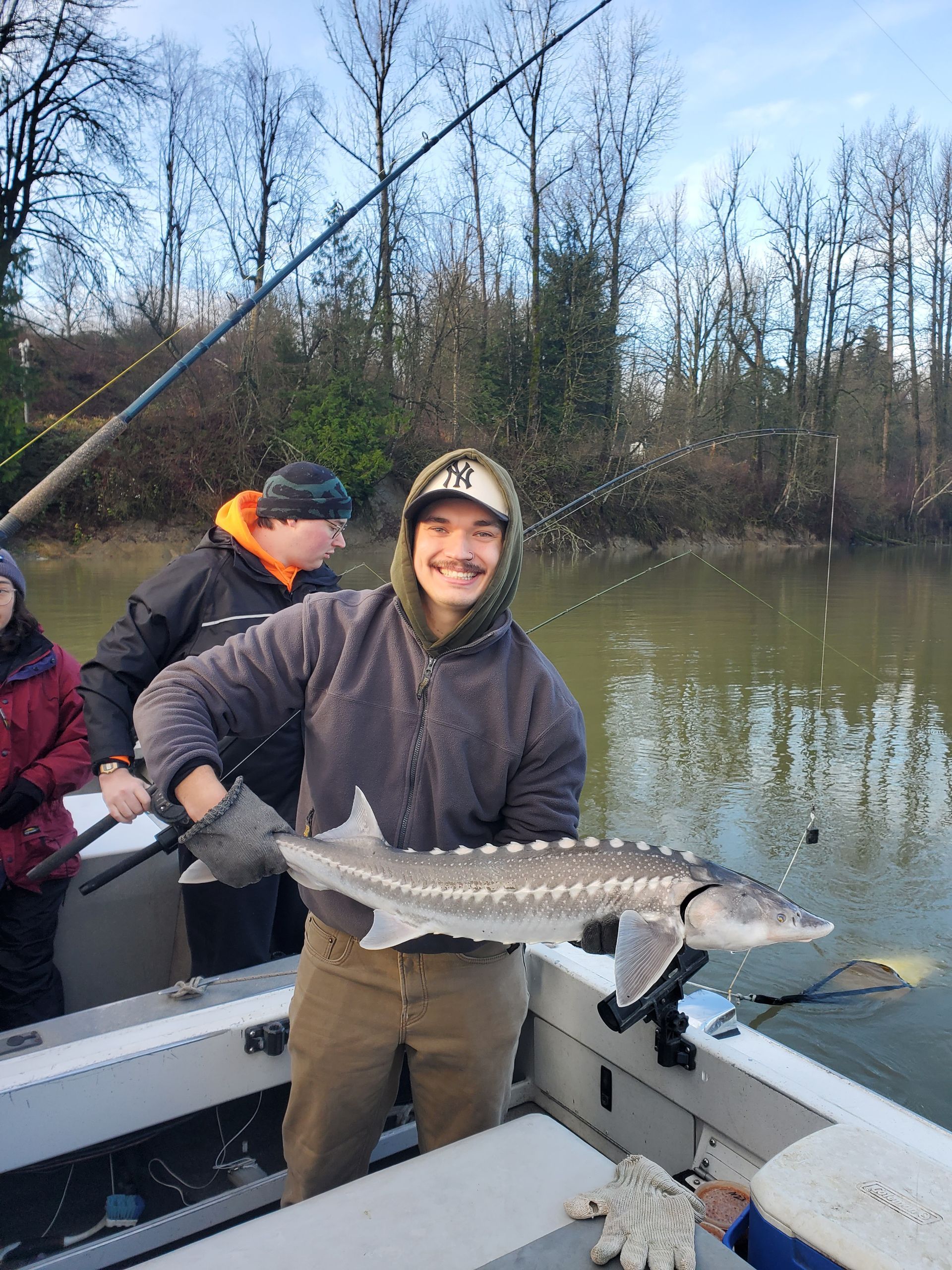 Person smiling on a boat, holding a large fish over the side with others nearby on a calm river.