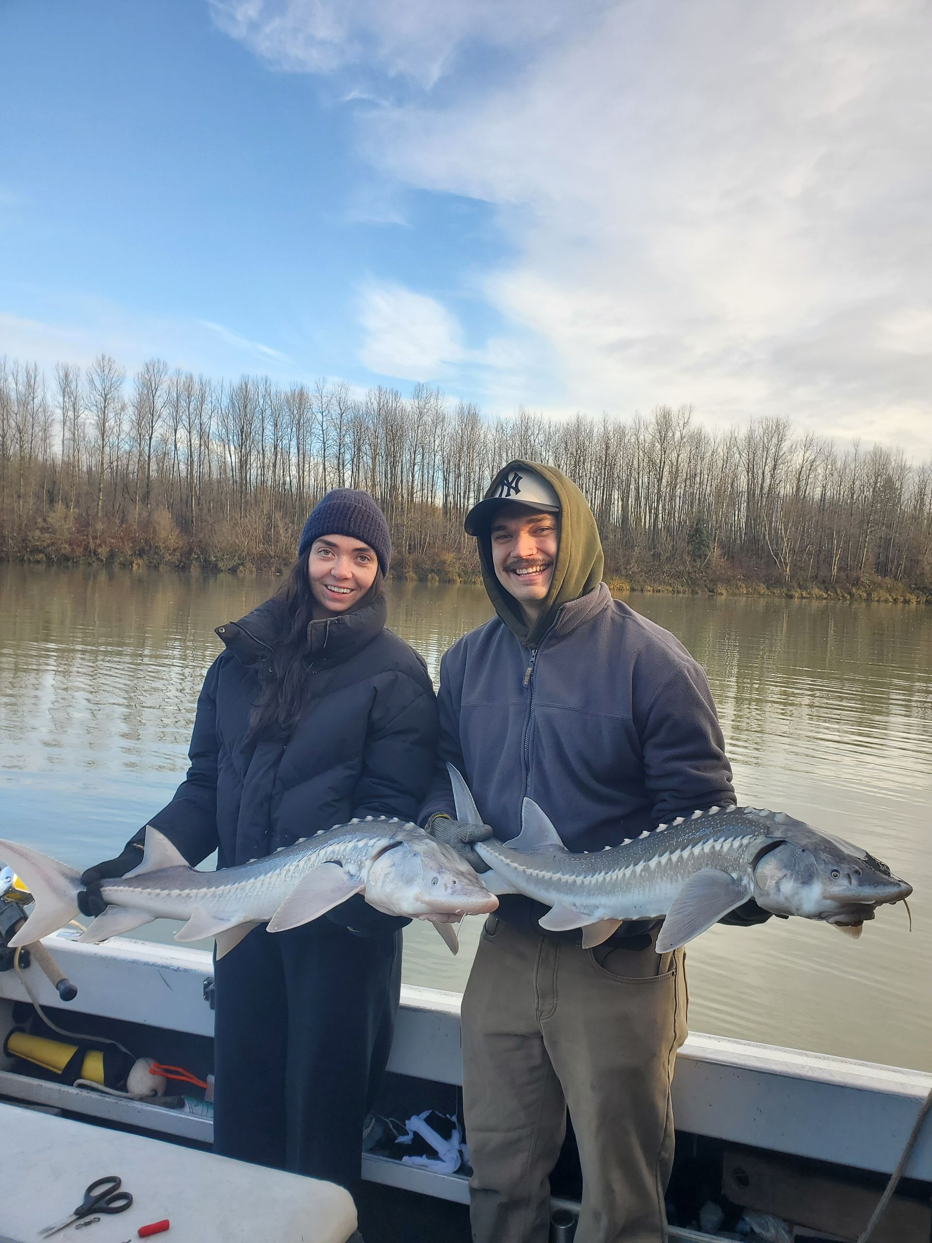 Two people on a boat holding large fish, with calm water and bare trees in the background.