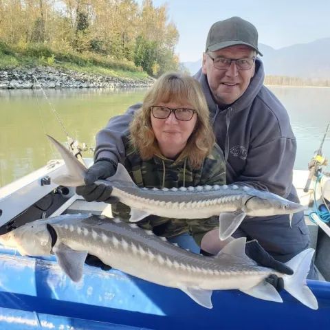Two people on a boat holding two large fish on a calm lake with mountains in the background