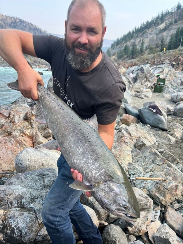 Smiling man holding a large silver fish on a rocky riverbank with mountains in the background