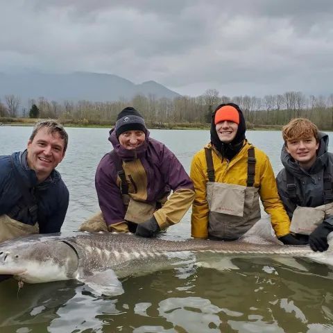 Four people posing with a large fish in a lake, wearing jackets and waders on a cloudy day.
