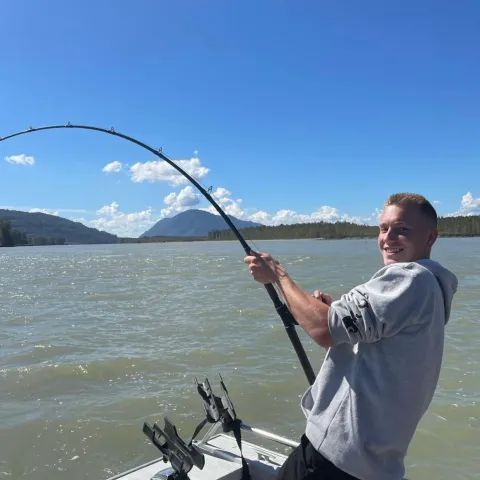 Person on a boat holding a bent fishing rod, smiling on a windy lake with mountains in the background