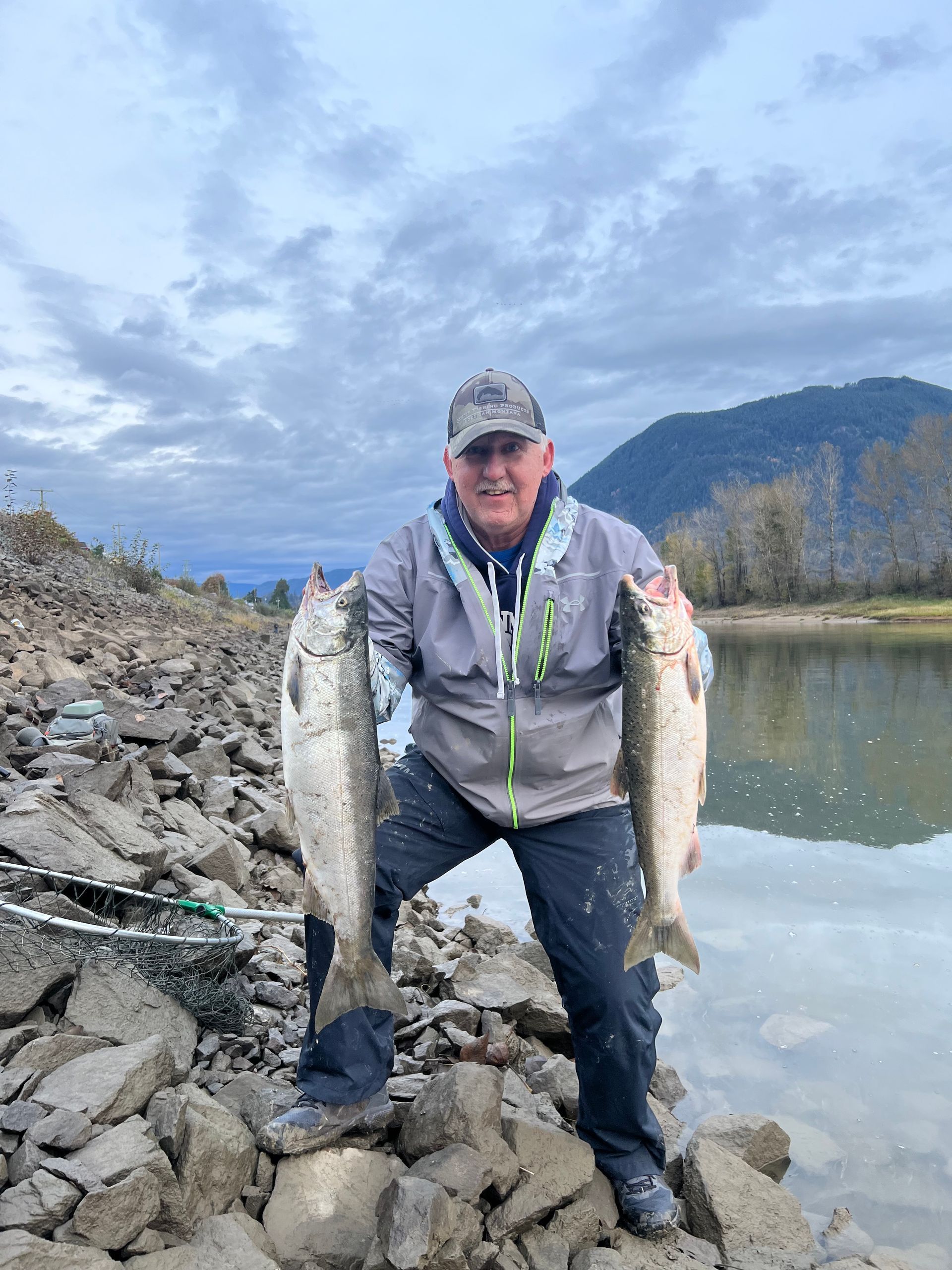 Person on a rocky riverbank holding two fish, with water, mountains, and cloudy sky in the background
