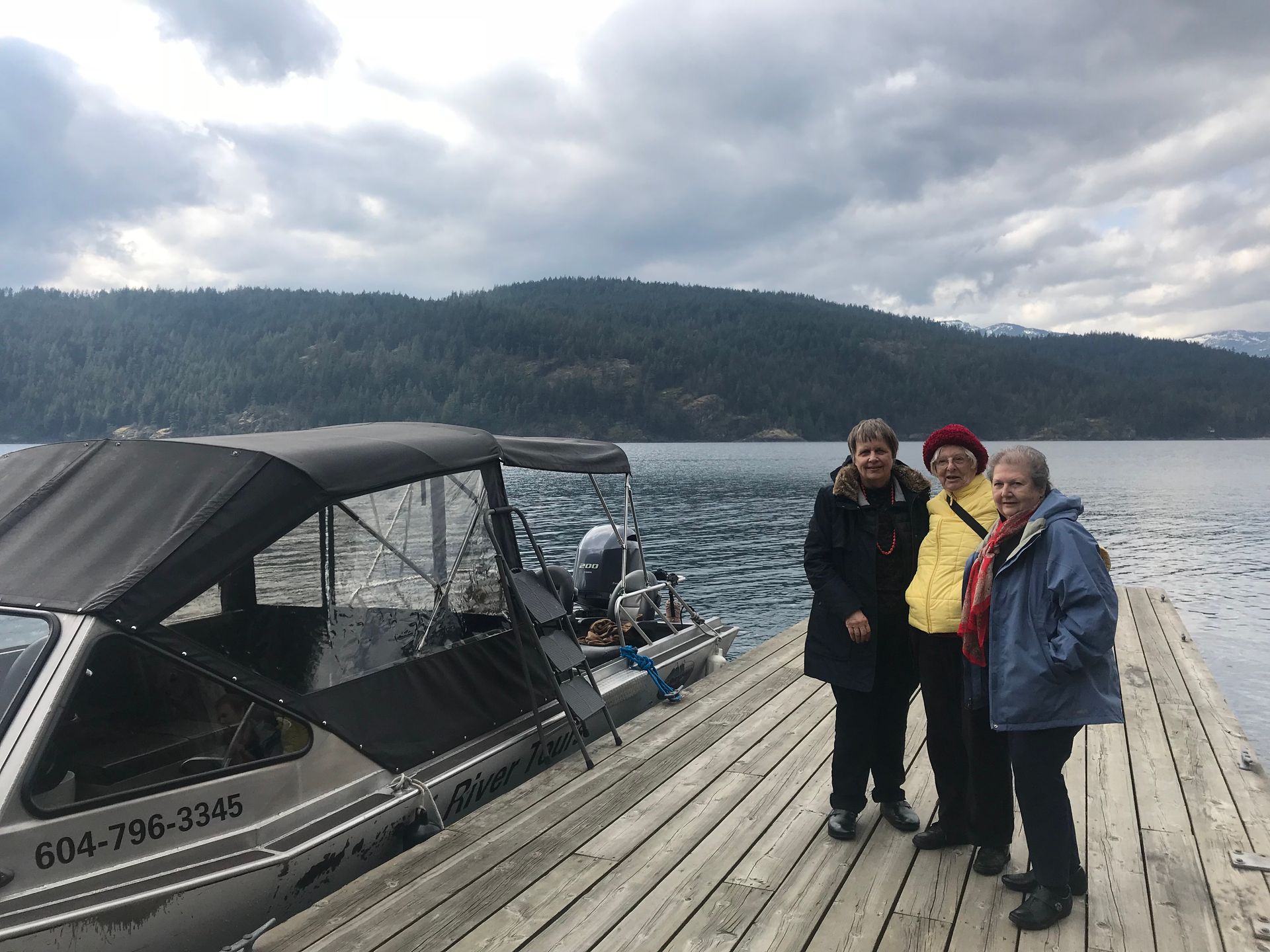 Three people on a wooden dock beside a covered boat, with choppy water and cloudy mountains behind.