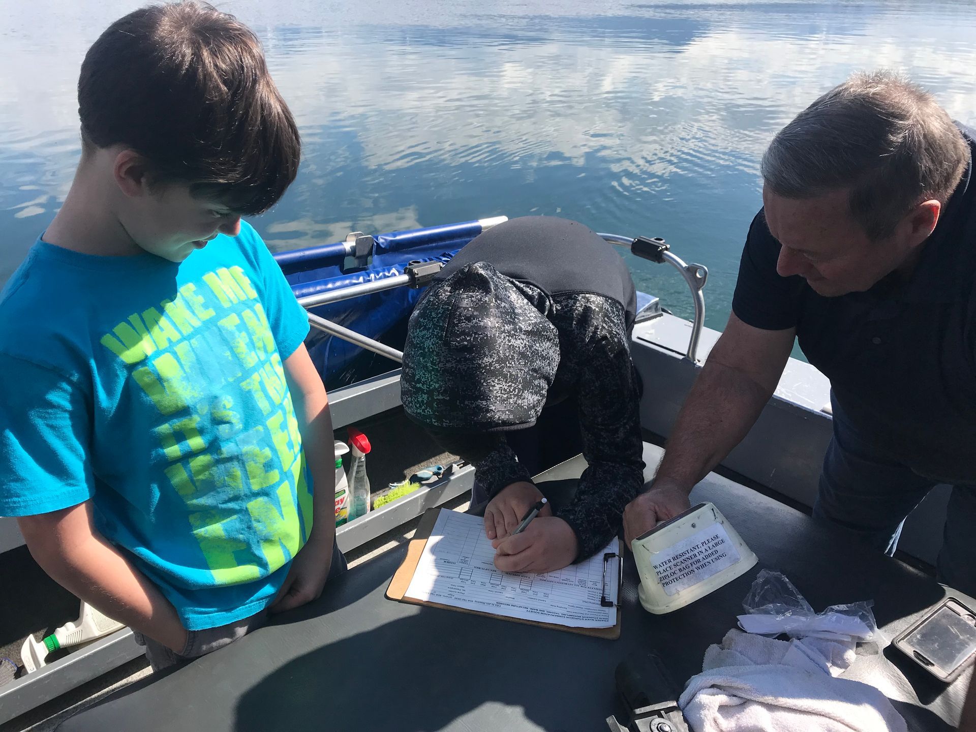 Two people on a boat near water, one writing on a clipboard while the other looks on.