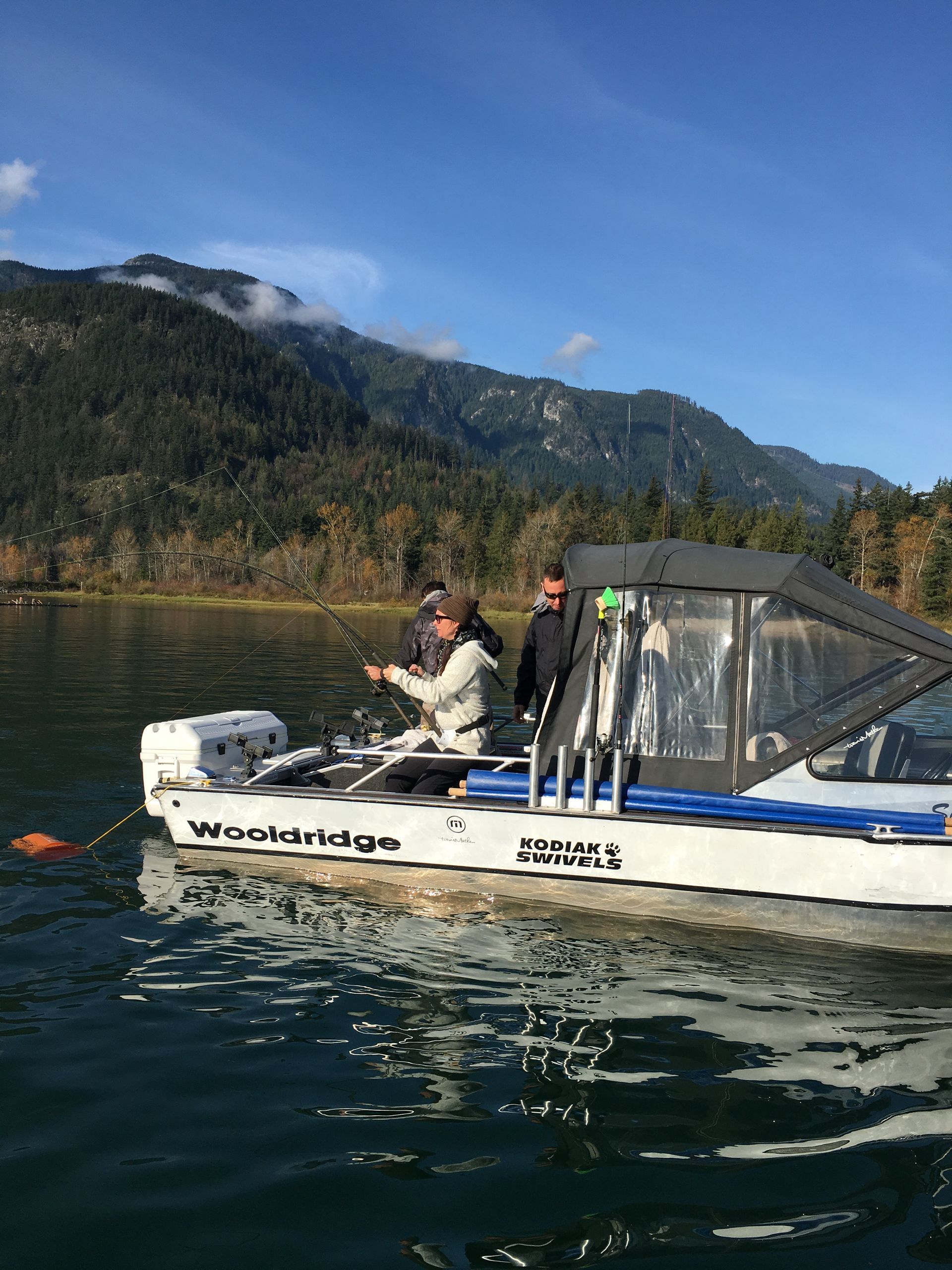 Two people fishing from a small white boat on a calm lake with mountains in the background