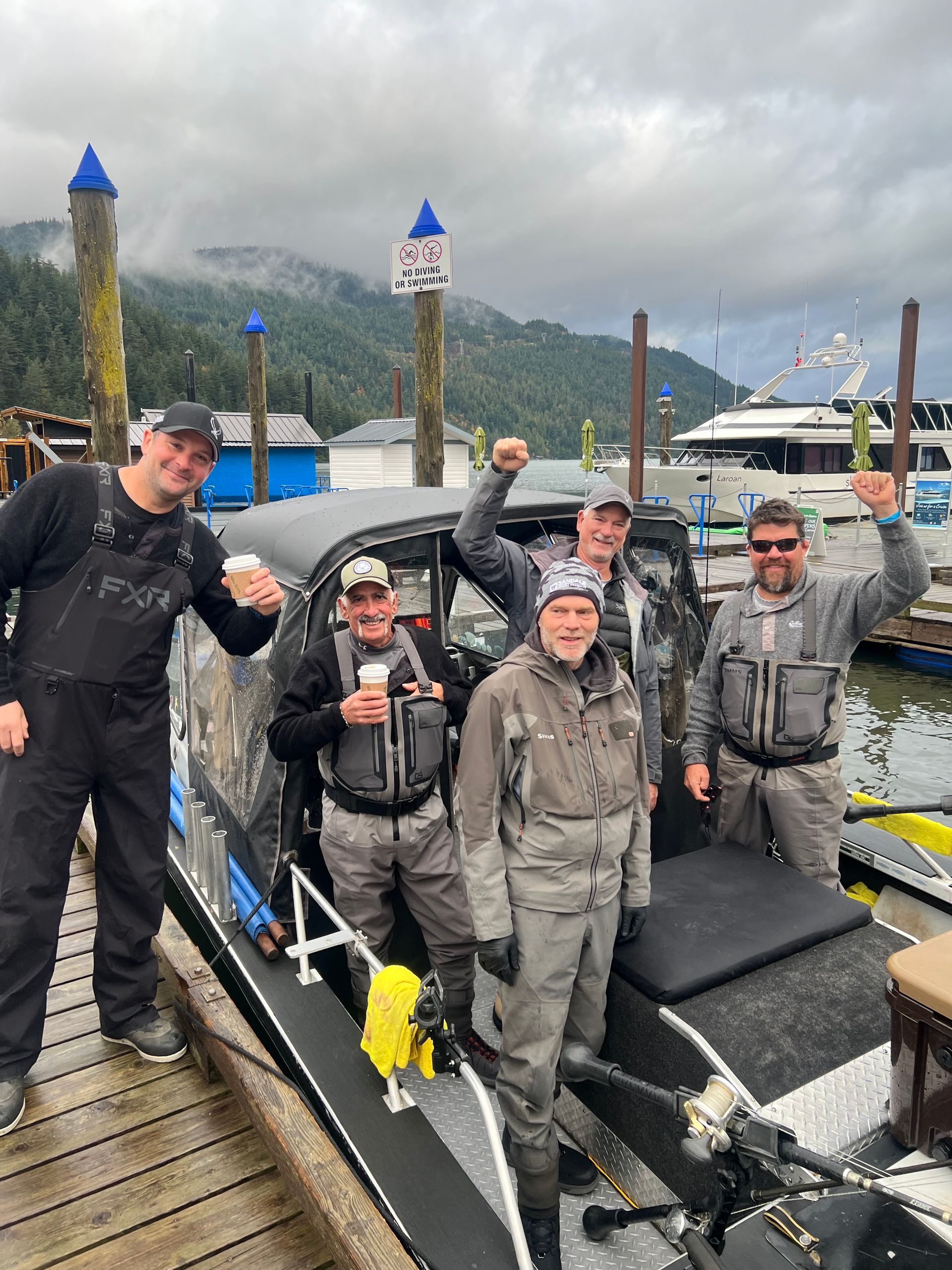 Five people in rain gear pose on a dock beside a boat in a cloudy marina.