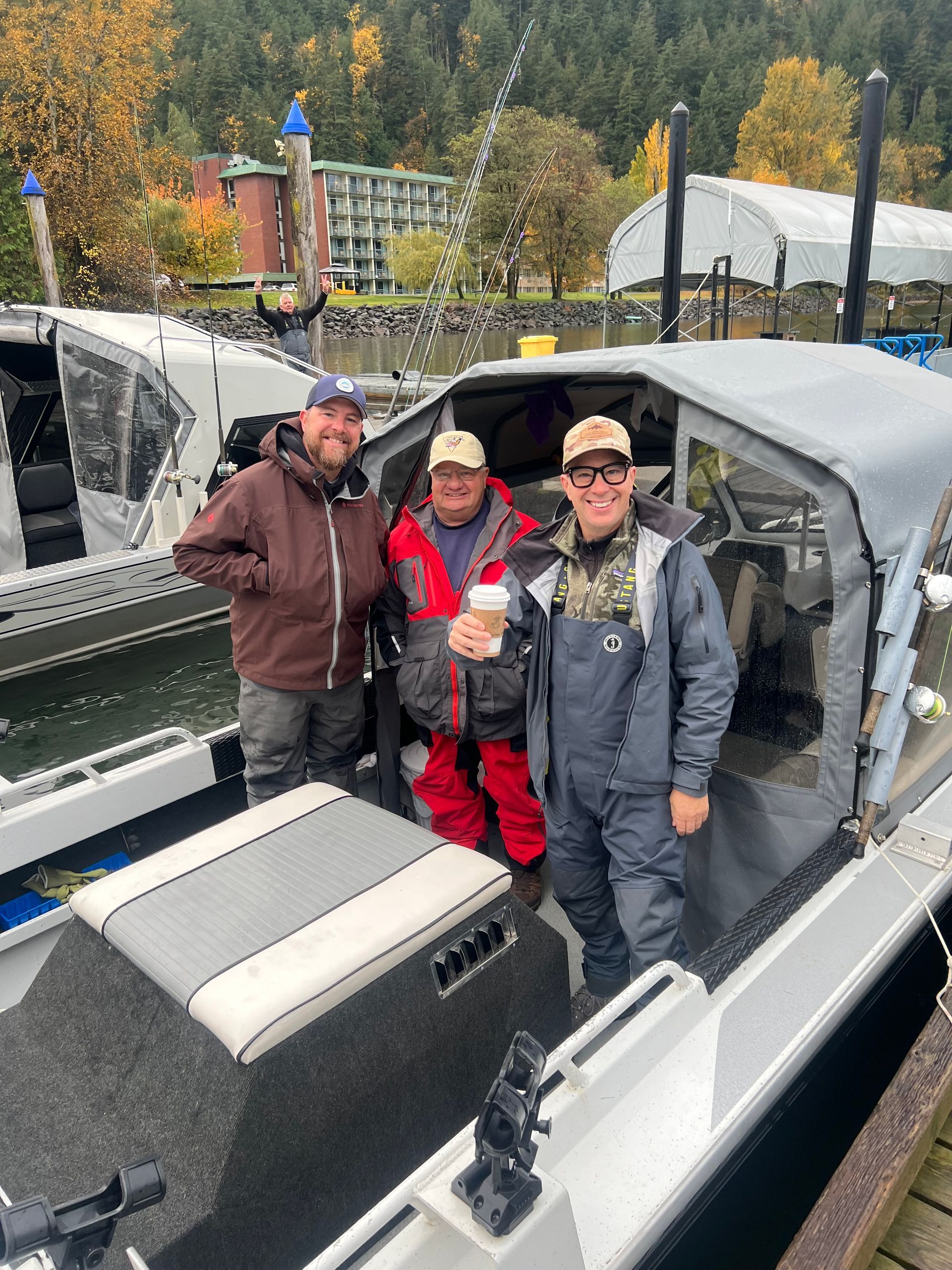 Three people posing in a boat on a canal, wearing jackets and hats, with autumn trees and buildings behind them.