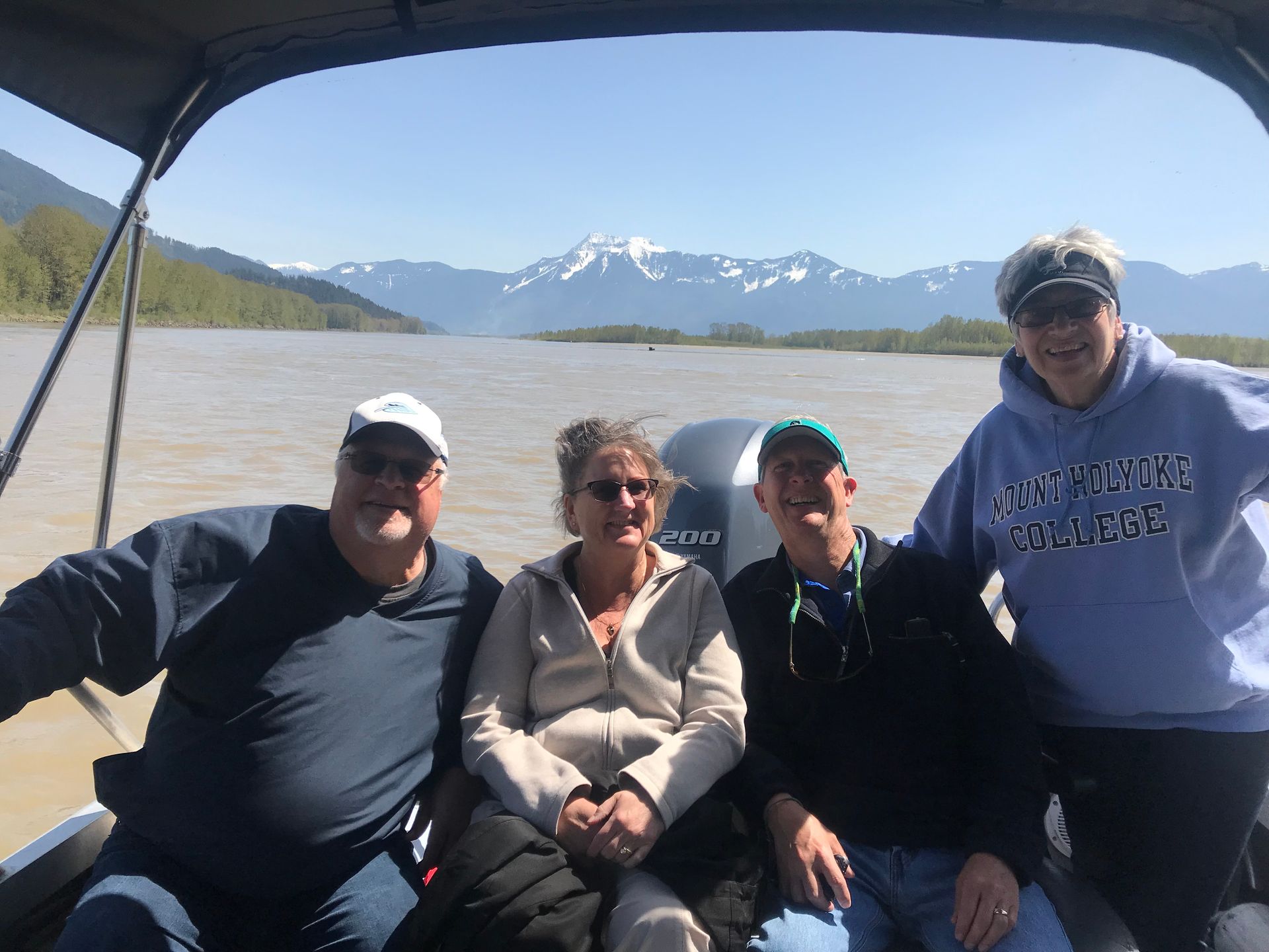 Four people smiling on a boat, with a lake and snowy mountains in the background.