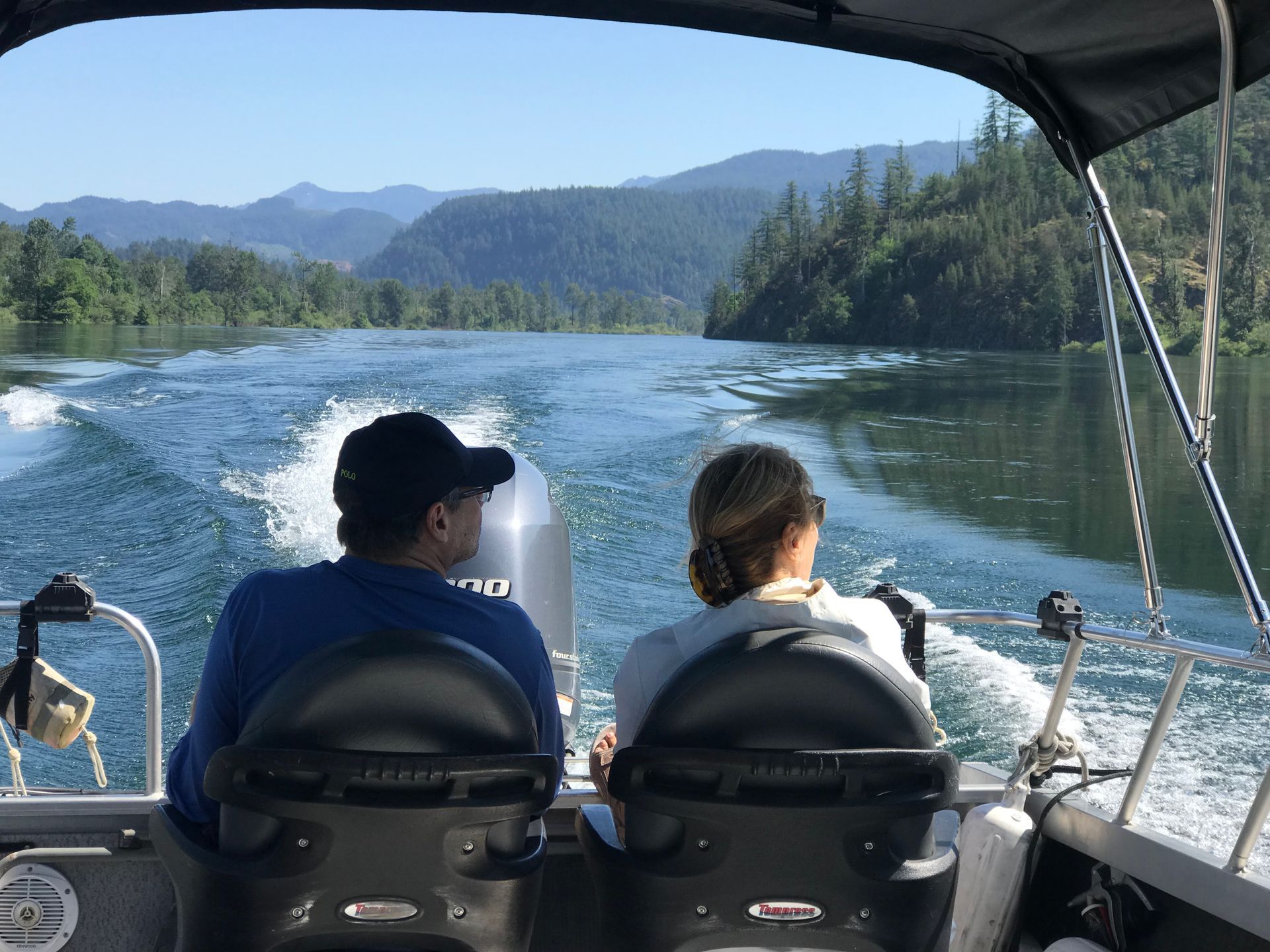 Two people riding in a motorboat on a lake, with forested mountains and a foamy wake behind them.