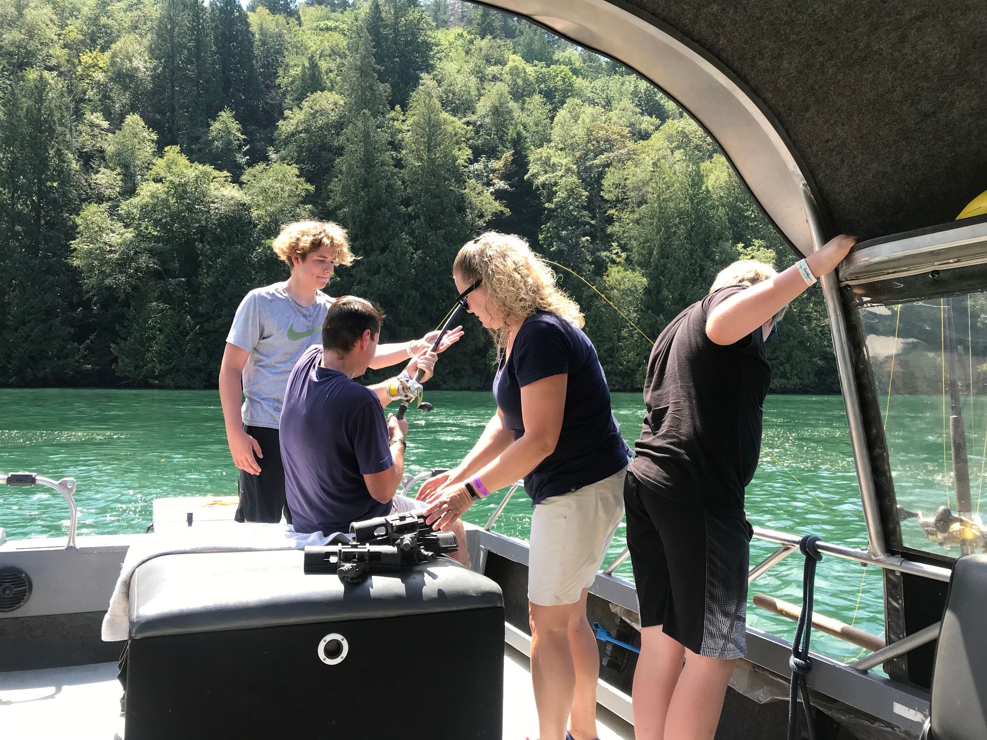 People on a boat deck by a green river, one pointing toward the water under a shaded canopy.