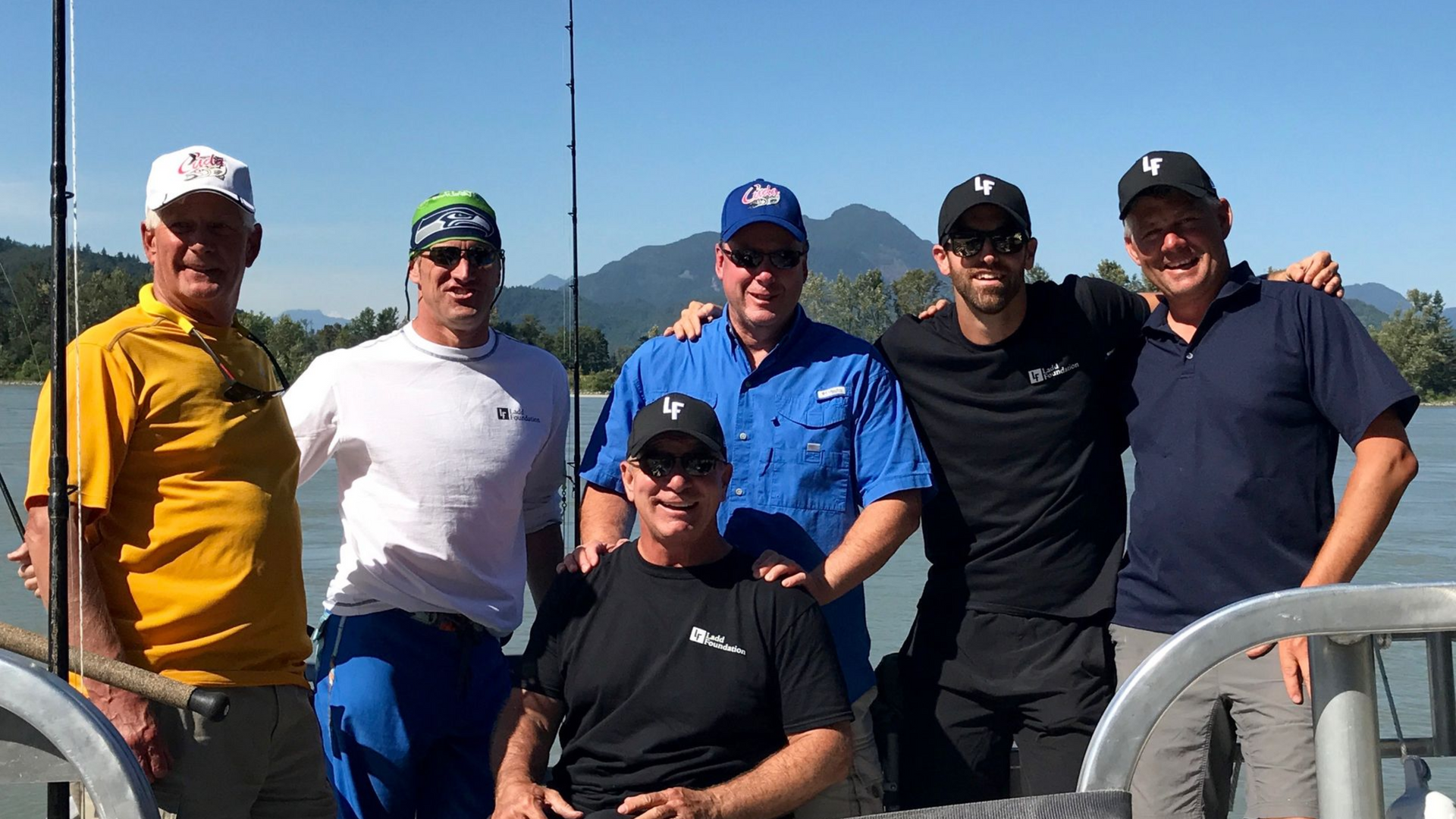 Six people posing on a boat deck with mountains in the background, smiling in bright daylight