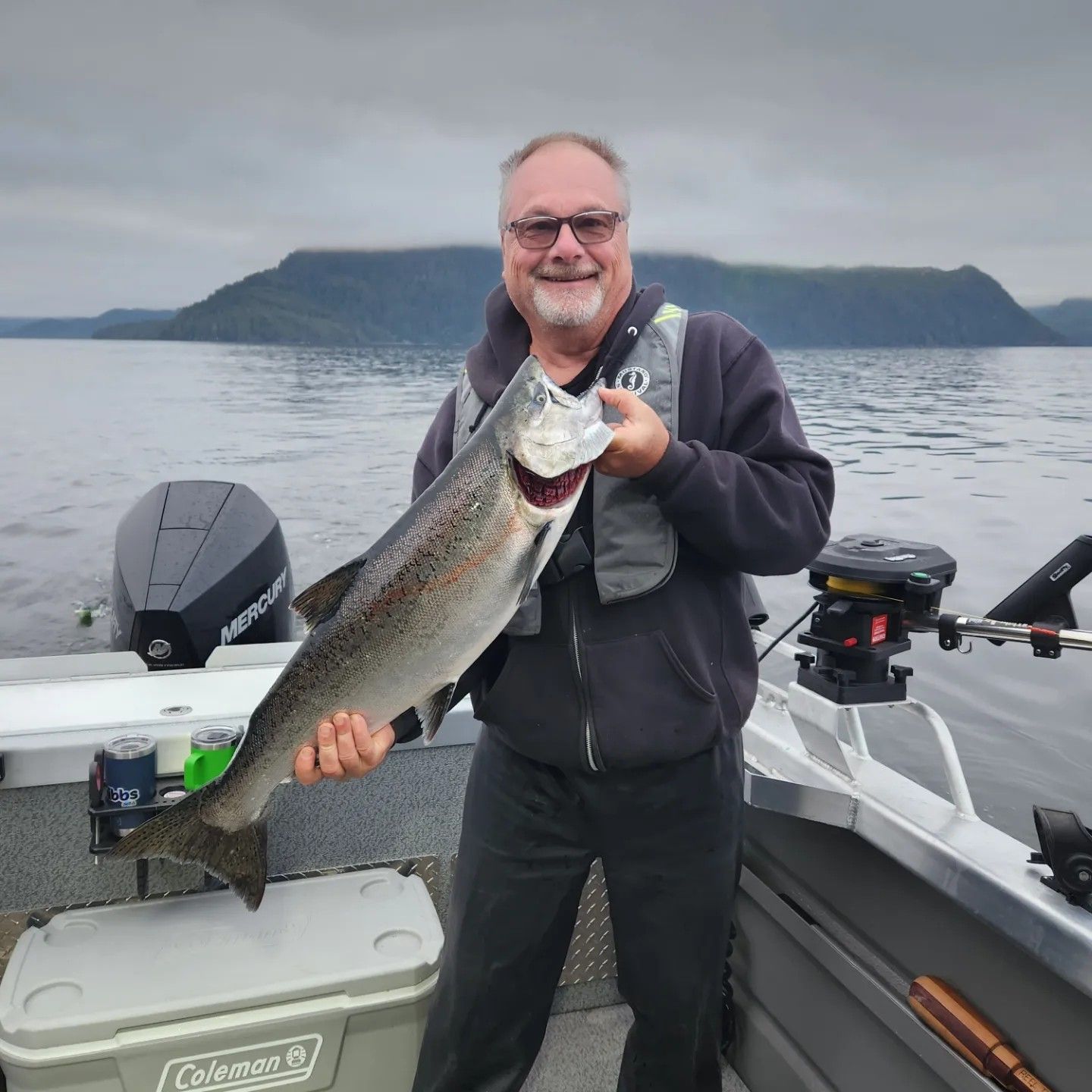 Man holding a large fish on a boat with a cloudy sea and mountain backdrop.