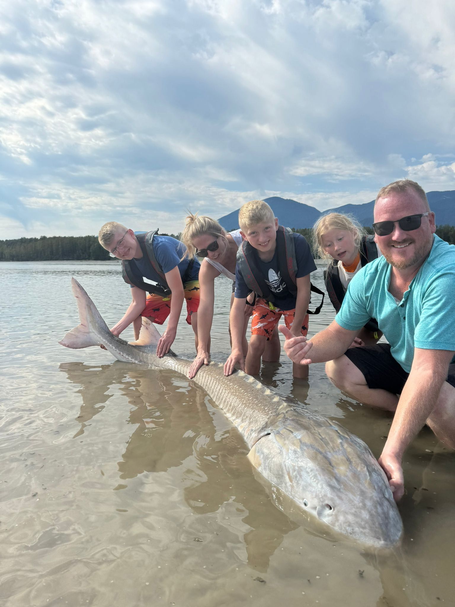 People kneeling beside a huge fish on a wet sandy beach under a cloudy sky