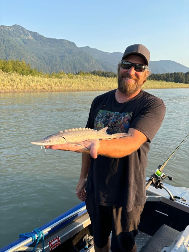 Man holding a large fish on a boat on a lake with mountains in the background