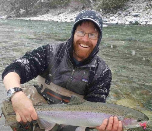 Smiling angler holding a large fish in a shallow river, wearing a hooded jacket and glasses.