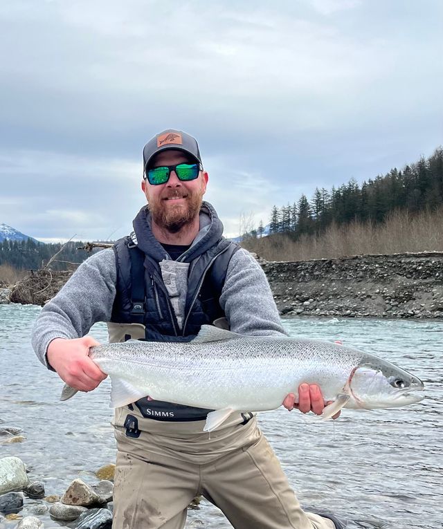 Angler holding a large silver fish in a river, with snowy banks and forested hills behind him.