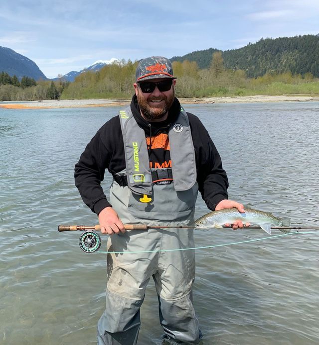 Smiling angler in waders holding a fishing rod in a river with mountains in the background