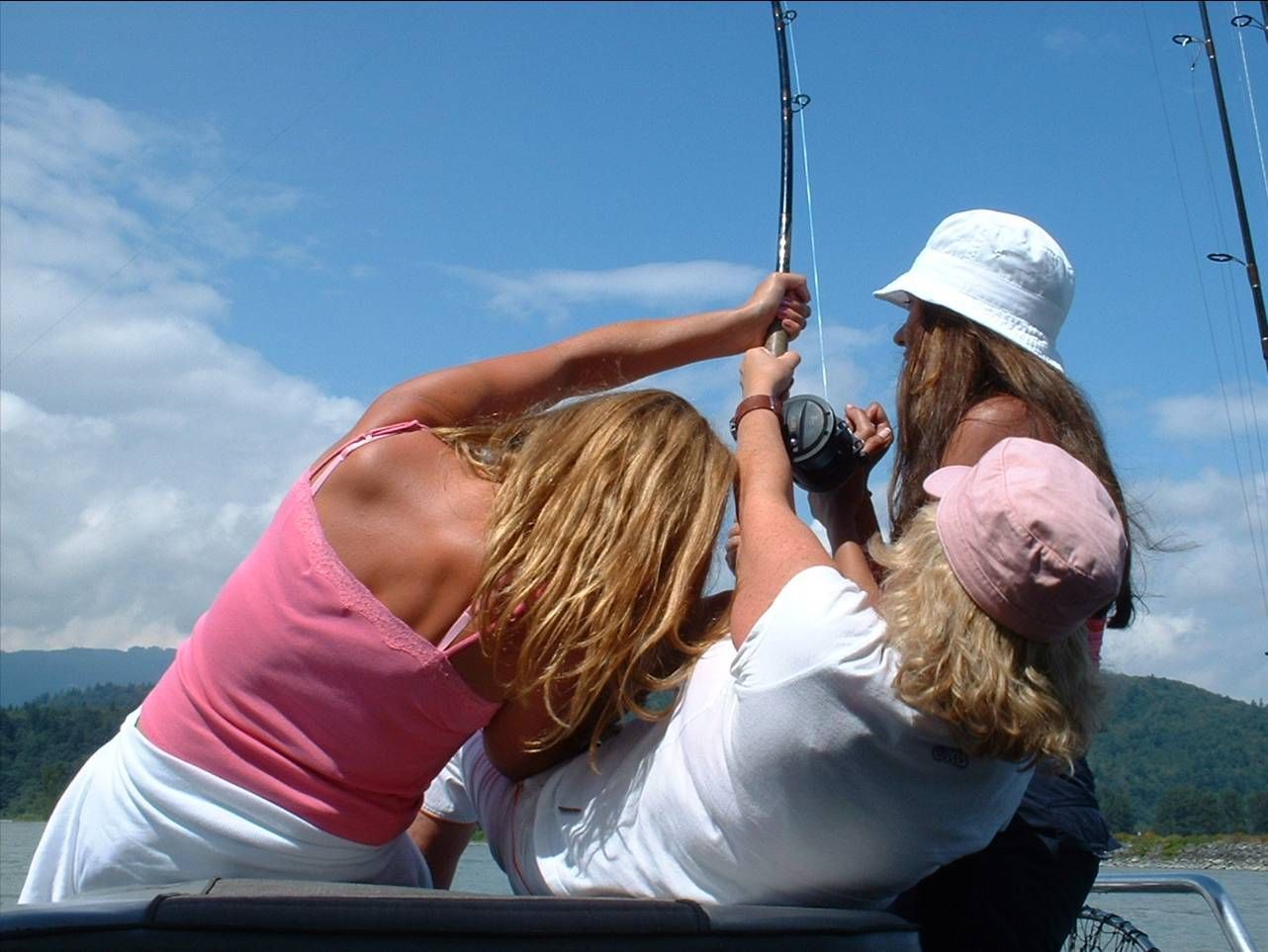 Three people on a boat hoisting a sail against a blue sky and distant green hills