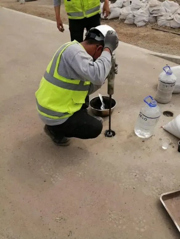 Un trabajador con un chaleco reflectante se arrodilla y taladra un suelo de hormigón junto a unos bidones de agua.