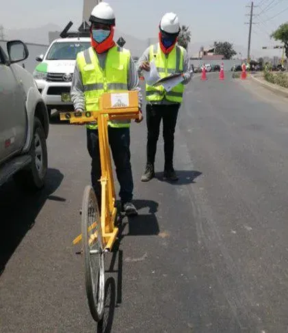 Dos operarios de carreteras, con chalecos de seguridad y mascarillas, inspeccionan una carretera utilizando una rueda de medición.
