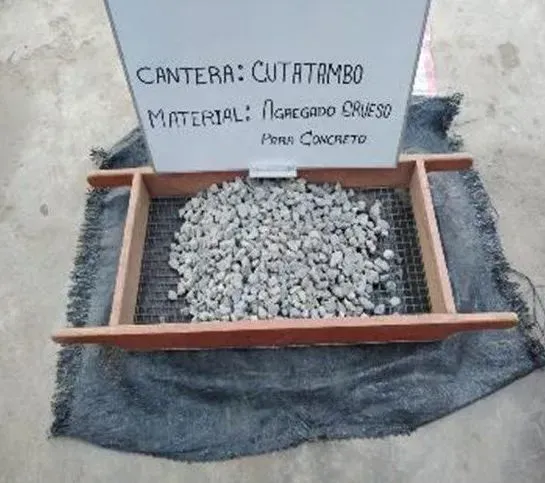 Pequeño marco de madera lleno de grava sobre una tela gris, con un letrero que dice “Cantera: cutrambó”.