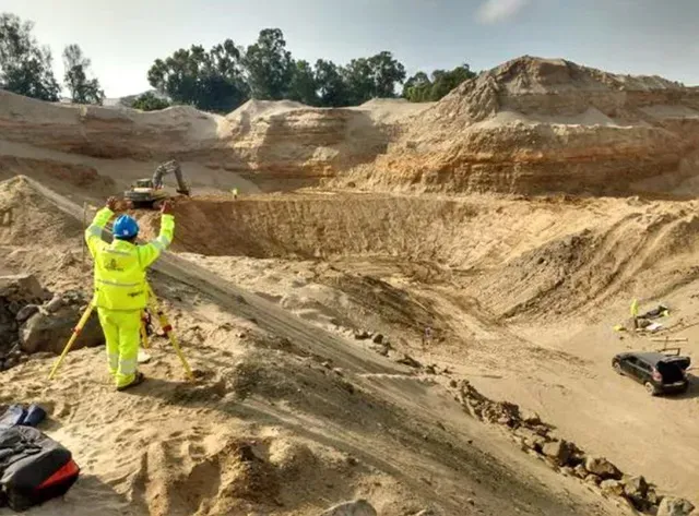 Un trabajador con equipo de seguridad de color neón inspecciona un sitio de excavación arenoso con taludes aterrazados.