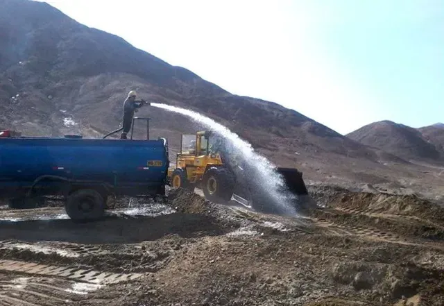Camión cisterna azul rociando agua en un camino de tierra polvoriento en una zona montañosa.