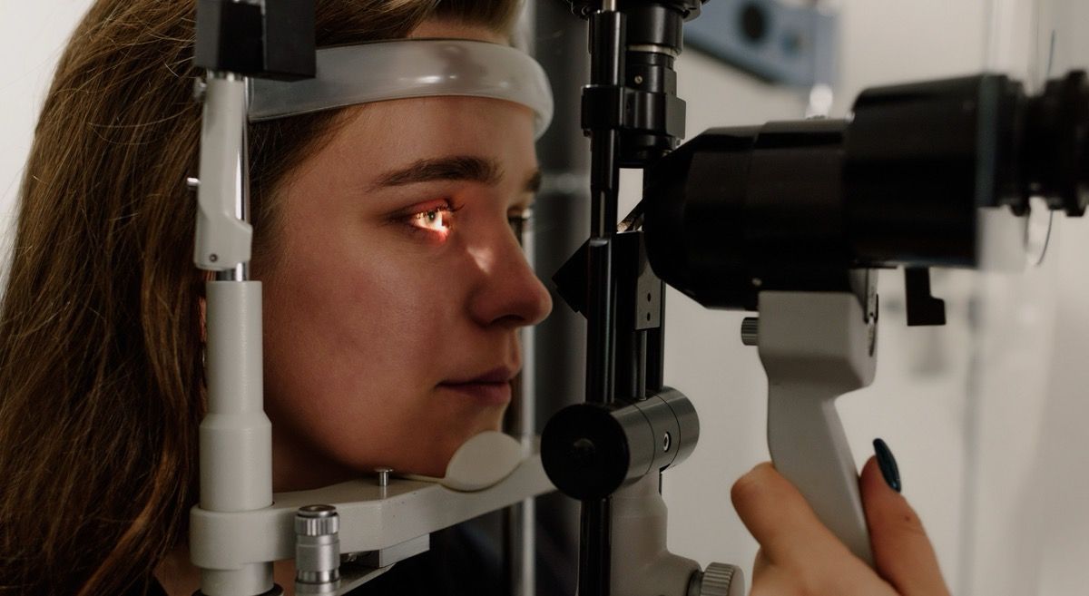 Person using a slit lamp eye exam machine, seen in profile, with chin resting on the device.