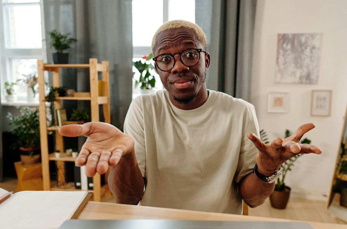 Man shrugging at a desk in a bright home office, with shelves, plants, and framed art in the background