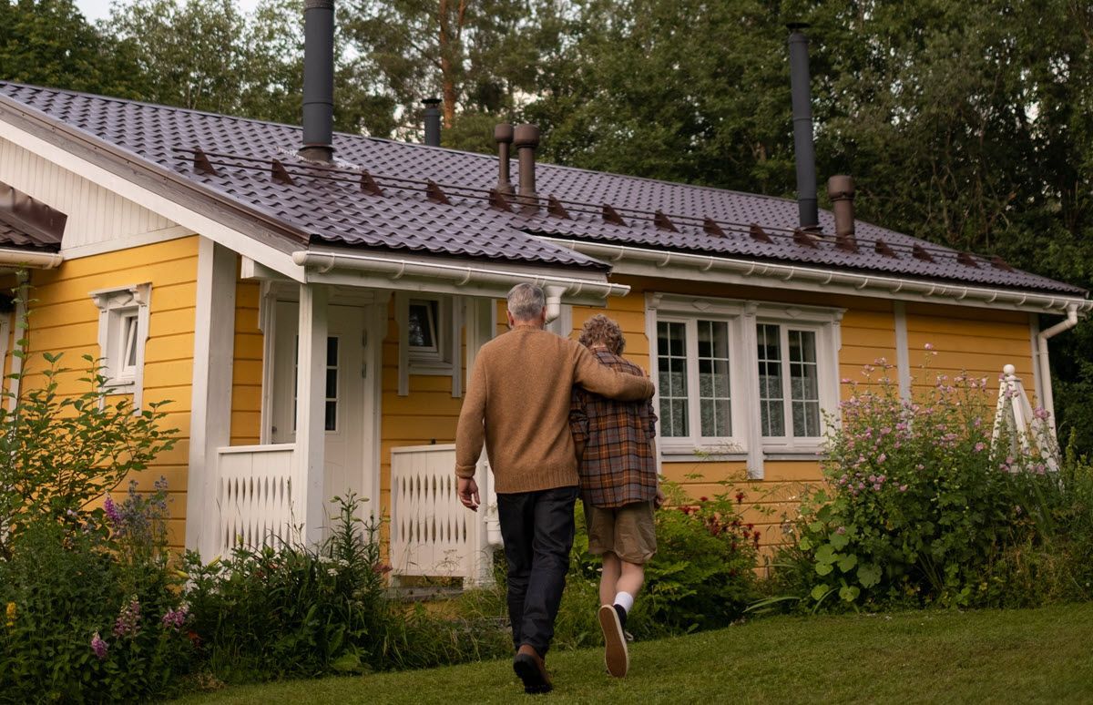 Two people walk arm in arm toward a yellow house with a white porch and wet roof.