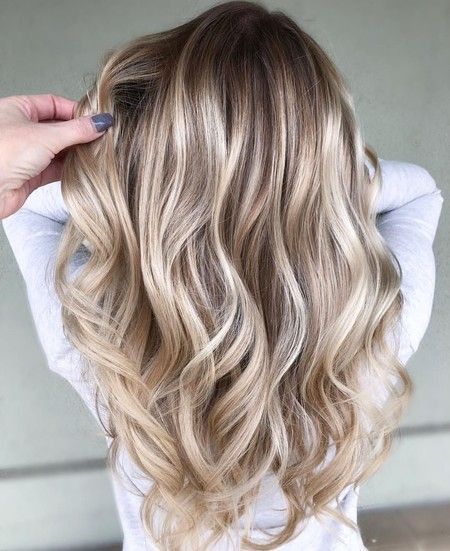 Una mano acaricia una larga melena ondulada con reflejos balayage rubios que se funden con una raíz más oscura.