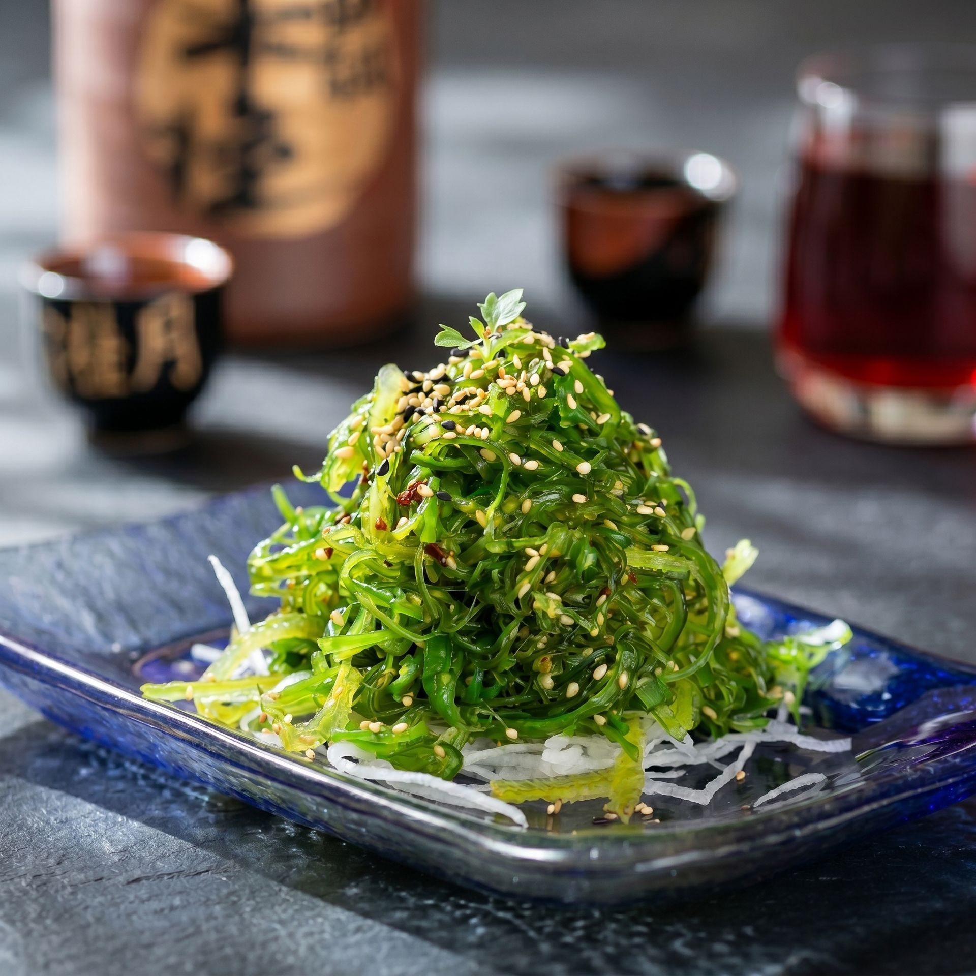 Green herb-topped food mound on a blue plate, garnished with sesame seeds on a dark table